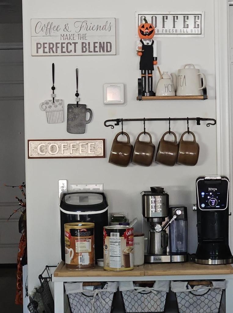 A kitchen coffee station featuring a decorative wall with signs about coffee and friendship, a pumpkin figure, and various coffee mugs hanging on a rack. The countertop displays coffee canisters, a coffee maker, and an ice maker.